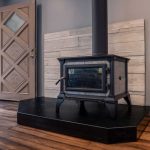 soapstone-hearth-wood-stove-door A soapstone hearth installed under a wood stove with a light-colored backsplash and brown, wooden floors in a contemporary home with an open door.
